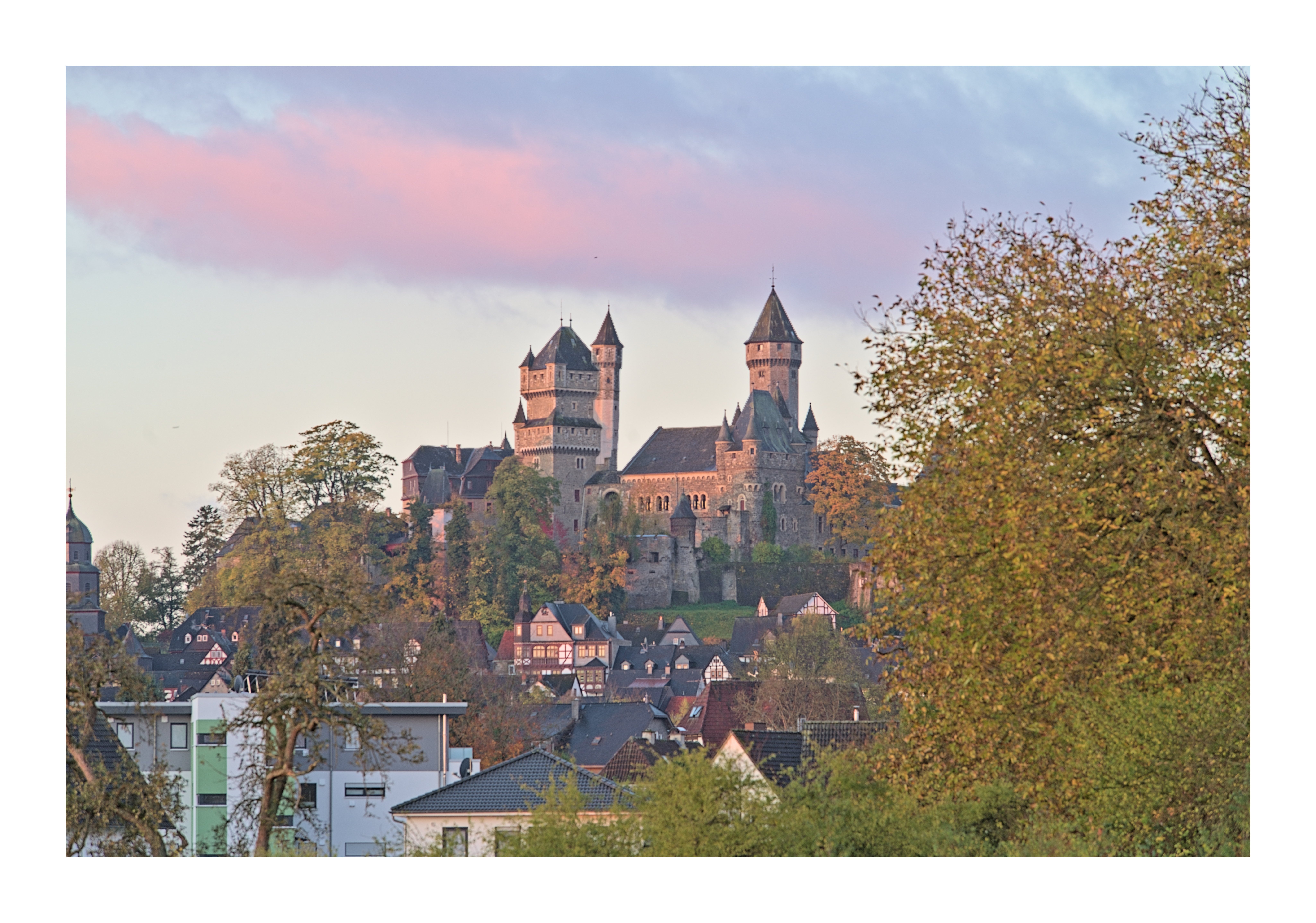 Das Bild zeigt die Burg Braunfels in den frühen Morgenstunden, eingebettet in eine ruhige und stimmungsvolle Landschaft...Die Burg thront majestätisch auf einem bewaldeten Hügel und wird vom sanften Licht der aufgehenden Sonne angestrahlt. Die warmen goldenen und rosafarbenen Töne des Morgenlichts verleihen den historischen Mauern und den markanten Türmen eine besonders eindrucksvolle Ausstrahlung. Über der Burg zieht sich ein zartes, rosa gefärbtes Wolkenband durch den Himmel, das den friedlichen Charakter der Szene unterstreicht...Am Fuße des Hügels schmiegen sich zahlreiche Fachwerkhäuser und Wohngebäude an den Hang und bilden den malerischen Ortskern von Braunfels. Im Vordergrund rahmen Bäume mit herbstlich gefärbtem Laub das Motiv ein und schaffen eine natürliche Kulisse, die den Blick auf die Burg lenkt...Insgesamt vermittelt das Bild eine ruhige, fast märchenhafte Atmosphäre und zeigt die Burg Braunfels als weithin sichtbares Wahrzeichen, das eindrucksvoll über der Stadt wacht und im weichen Morgenlicht besonders imposant erscheint.